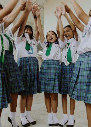 A group of joyful schoolgirls in uniforms raising their hands in celebration indoors.